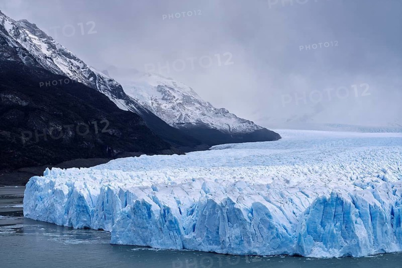 Perito Moreno glacier with shimmering blue ice break edge in Lake Argentino, snow-capped mountains under cloudy sky in the background, Los Glaciares National Park, Santa Cruz Province, Patagonia, Argentina