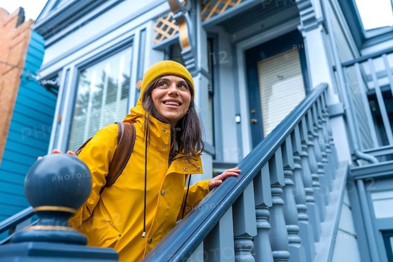 Woman wearing yellow raincoat and beanie smiling while ascending the exterior stairs of a classic victorian house in san francisco, capturing the essence of urban exploration and travel lifestyle