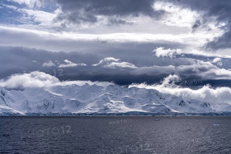 Snowy mountain coast with glacier edge at sea edge under dramatically cloudy sky, Antarctic Peninsula, Antarctic