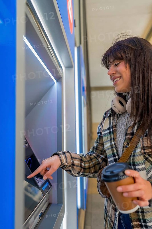 Smiling young woman engaging with an automated teller machine, holding a takeaway coffee cup, performing a financial transaction in an urban environment