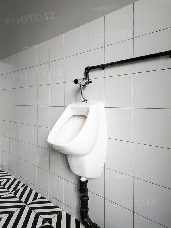 A urinal is mounted on a wall in a restroom. The tiles are white, and the floor has a pattern of black and white stripes. There is a pipe above the urinal connected to the plumbing