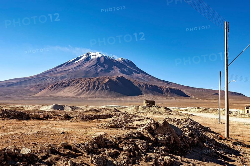 Large, snow-covered mountain above a barren, rocky landscape under clear skies, the town of Ollagüe in the Chilean Atacama Desert with the Ollagüe volcano of the same name in the background