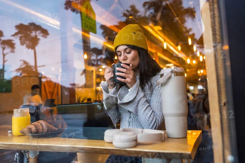 Young woman wearing a beanie enjoying a warm drink and breakfast at a wooden counter inside a coffee shop, with city reflections on the window and blurred street lights outside