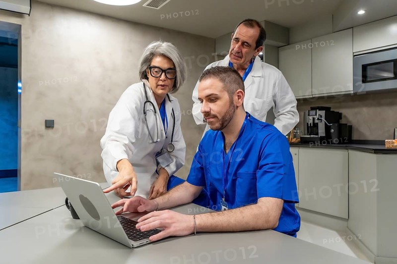 Medical professionals in scrubs and lab coats collaborating on a laptop, sharing information, and discussing healthcare data in a modern office or staff room