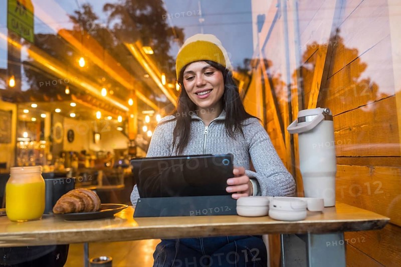 Smiling woman sitting at a table in a bustling san francisco coffee shop, happily interacting with her tablet while enjoying a croissant and orange juice, embodying modern digital nomad lifestyle
