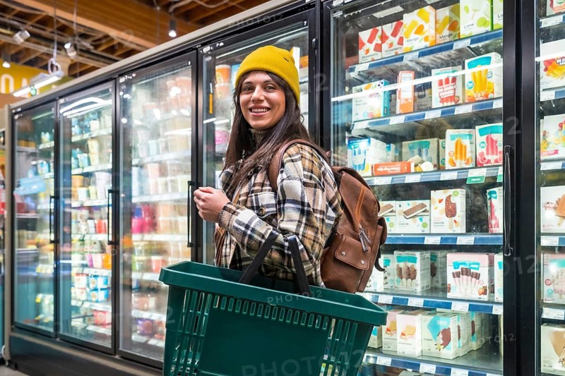 Young woman shopping for groceries in a supermarket aisle, smiling and holding a shopping basket while looking at frozen food and ice cream products in refrigerated display cases