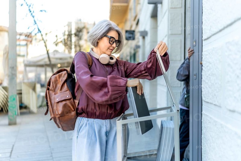 Stylish mature woman with gray hair and glasses returning a magazine rack to a doorway, performing a daily task in an urban environment while commuting