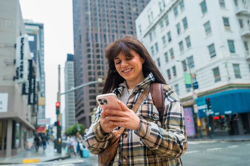 Young woman smiling, happily using her mobile phone for communication and social media, walking along a san francisco urban street with office buildings