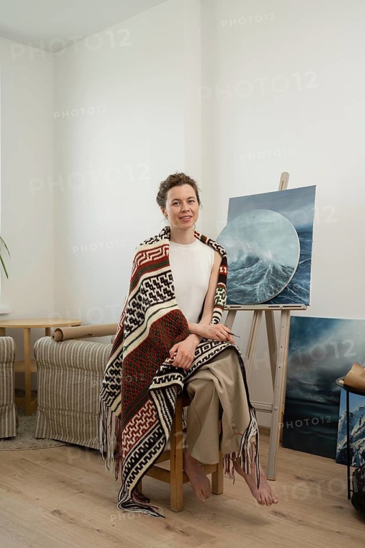 An artist sits barefoot in a cozy home studio, draped in a colorful patterned blanket. She is surrounded by ocean themed canvas paintings, exuding creativity and tranquility