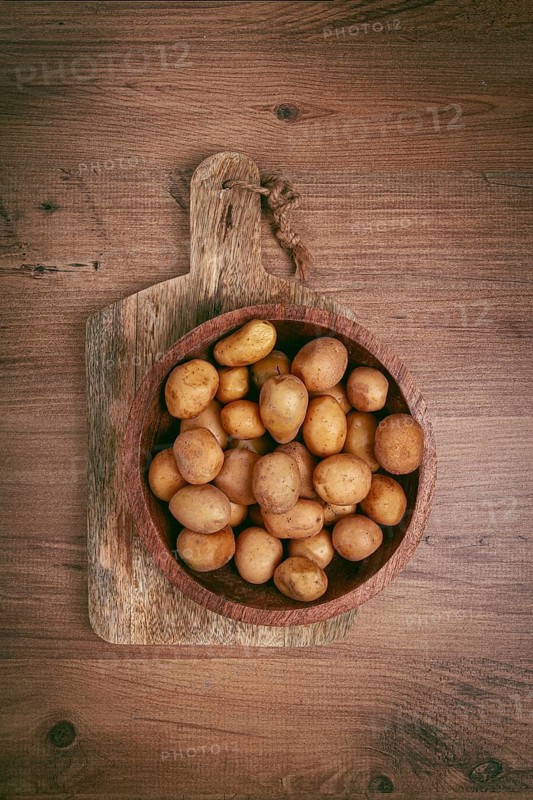 A collection of small, golden-brown potatoes fills a rustic wooden bowl placed on a textured wooden surface, emphasizing their natural appeal and freshness
