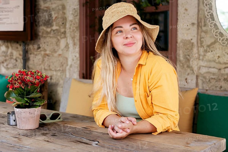 A young woman in a vibrant yellow shirt and sun hat sits at a rustic cafe table with a potted plant, enjoying her surroundings and looking away thoughtfully