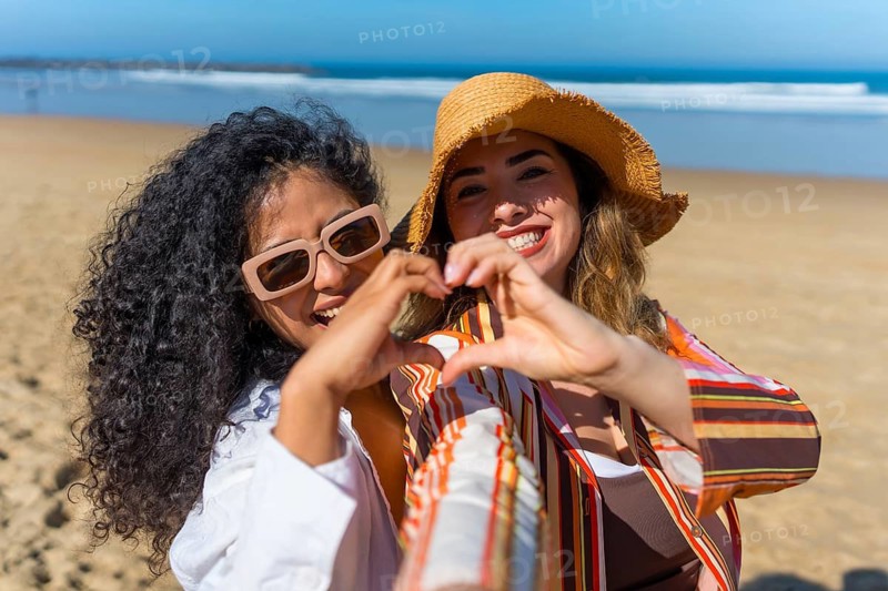Two smiling young women celebrating their friendship and loving summer while doing a heart symbol with their hands in front of the ocean on a sunny day at the beach