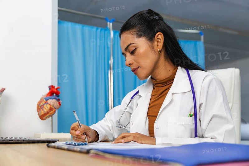 A dedicated doctor in a white coat writes notes at a desk in a medical office. A heart model is visible, signifying healthcare and professional commitment in a clinical setting