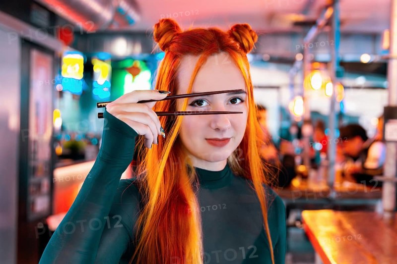 A young woman with vibrant hair and a stylish outfit enjoys her time at an Asian food market, playfully holding chopsticks The colorful, lively market atmosphere surrounds her