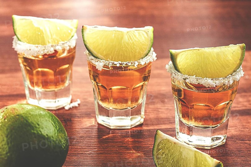 Three glasses with tequila and salt on the rims are arranged on a wooden table. Each glass has a slice of lime on top. A lime sits in the background under natural light