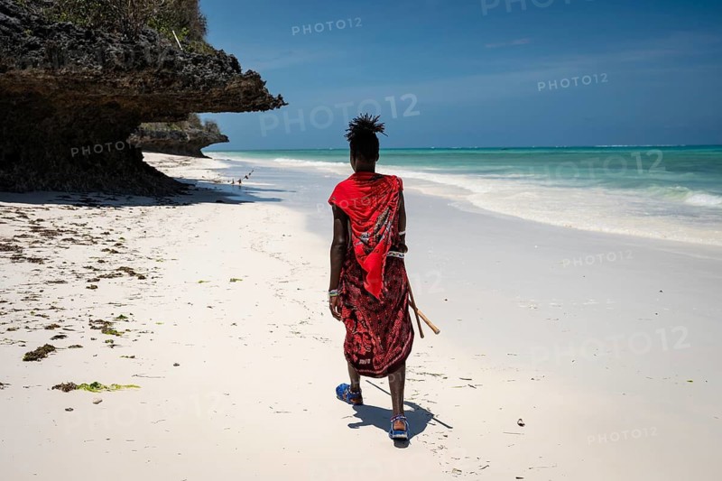 A Maasai man in traditional red attire walks away from the camera along a pristine white sand beach, next to a dramatic coral rock overhang. In Zanzibar, Tanzania