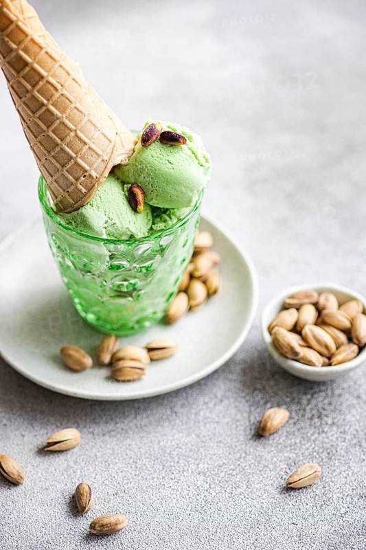 A close up of pistachio ice cream scoops in a green glass with a waffle cone, garnished with pistachios. Served on a plate with scattered pistachios for decoration