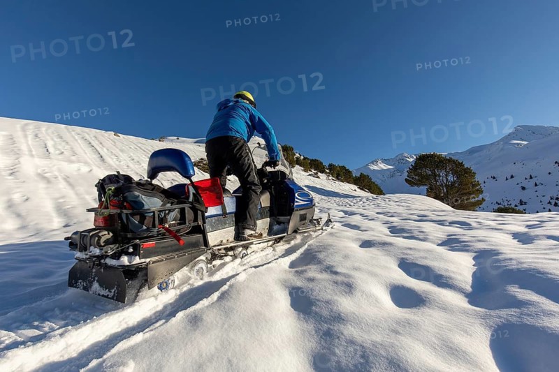 A snowmobiler in blue jacket navigates snowy terrain under a clear winter sky. The sun casts long shadows on untouched snow, capturing the thrill of outdoor adventure