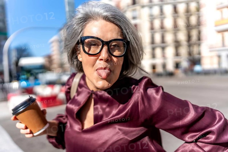 Playful woman expressing humor with her tongue out, holding a coffee cup and wearing eyeglasses on a sunny urban street, embodying vibrant individuality and joy