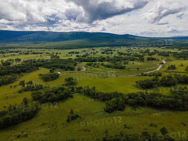 Vast green fields stretch across Vladivostok, Russia, under a cloudy sky. A winding river cuts through the landscape, surrounded by lush vegetation. This scene captures the tranquility and beauty of the region's natural environment