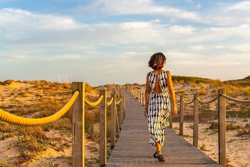Woman walking on a wooden boardwalk, surrounded by golden sand dunes and dry grass, heading towards the horizon during a warm sunset in seignosse, france, enjoying vacation
