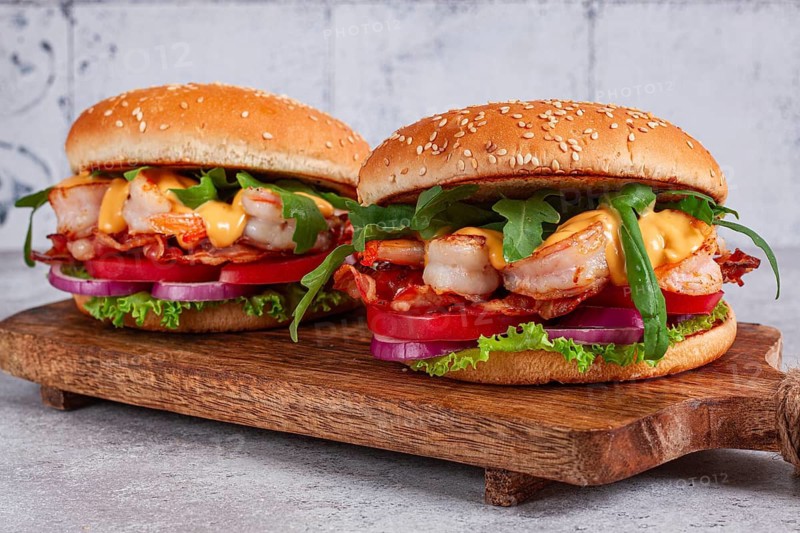Two shrimp burgers and buns with sesame seeds, bacon, lettuce, tomatoes and onions, On a wooden board