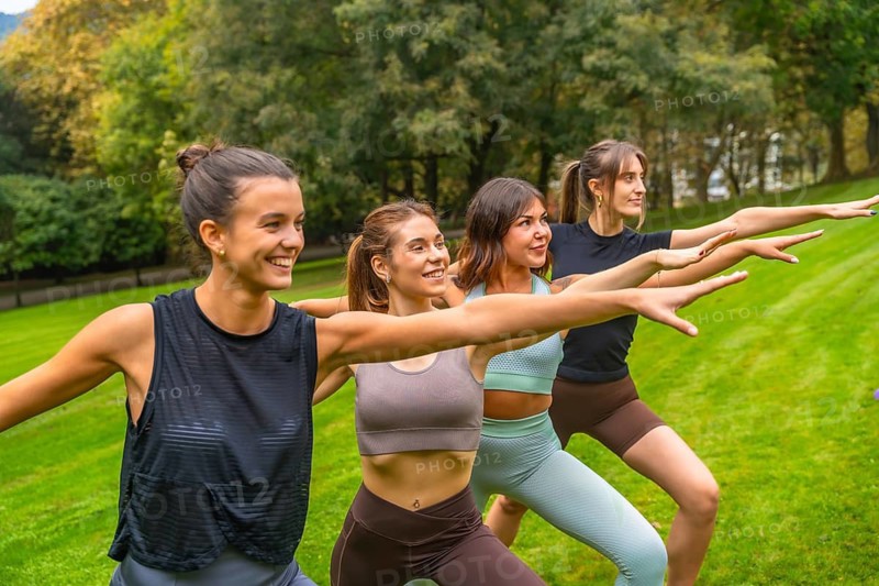 Group of happy young women practicing warrior pose an outdoor yoga session, promoting wellness, friendship, healthy lifestyle, and mindful exercise in a lush green park