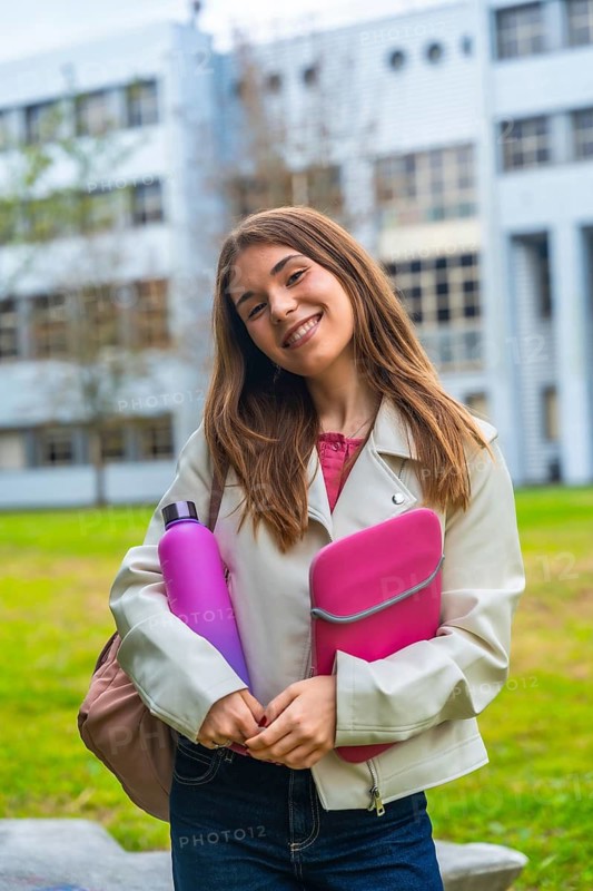 Young female college student smiling at camera, standing on campus with a laptop sleeve and reusable water bottle, representing education and sustainability