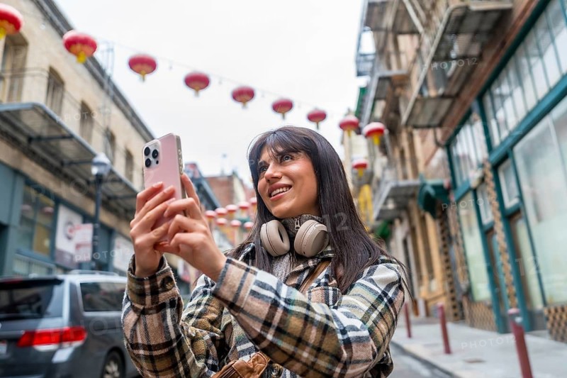 Young woman smiling and holding a smartphone, capturing content while exploring the vibrant street culture of san francisco's chinatown, amidst traditional red lanterns and historic buildings