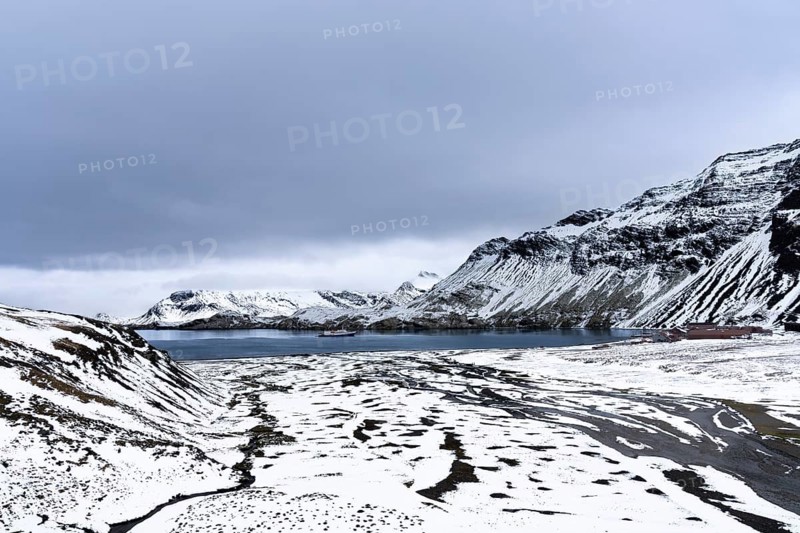 Snow-covered bay with expedition ship and former whaling station on the shore, surrounding mountains under overcast sky, Stromness Bay, South Georgia