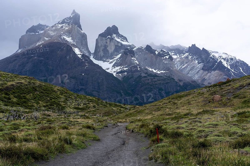 Cuernos del Paine, snow-capped granite peaks under cloudy sky, hiking trail with red marking leads through grassy hills, Torres del Paine National Park, Patagonia, Chile