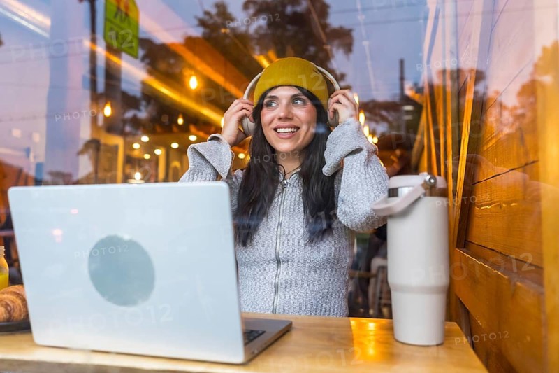 Young woman confidently adjusting headphones while sitting at a coffee shop window with a laptop and a reusable mug, enjoying an urban remote work experience