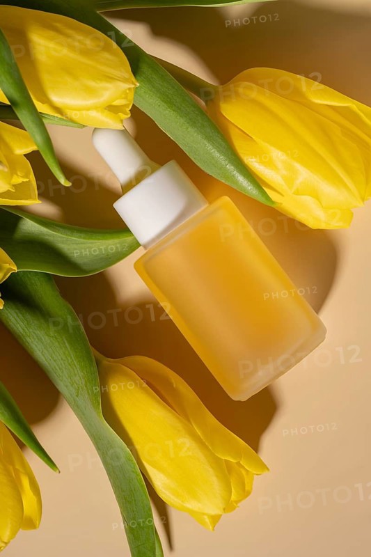 A flat lay composition featuring a frosted dropper cosmetic of facial serum surrounded by vibrant yellow tulips on a warm, neutral background. Perfect for skincare and beauty themes