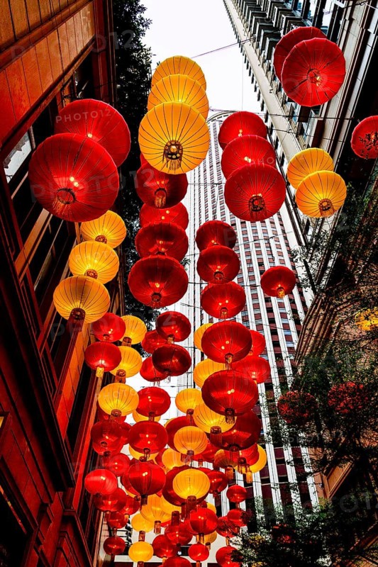 From below radiant red and yellow lanterns hang between skyscrapers in Hong Kong, celebrating Chinese New Year. The festive decorations create a vibrant canopy against the urban backdrop, symbolizing prosperity and good fortune