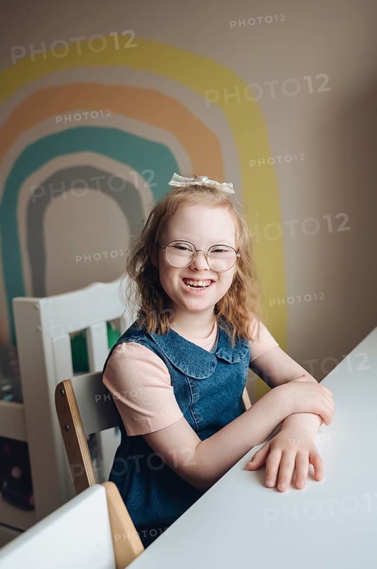 A joyful young girl with Down syndrome sits at a table in a colorful, playful room. She is smiling warmly, radiating happiness and positivity, surrounded by vibrant decor