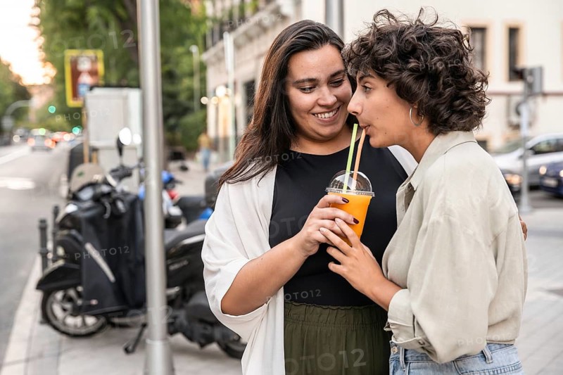A lesbian couple shares a refreshing drink with two straws, smiling lovingly at each other, while standing on a bustling urban street, capturing a moment of joy and connection