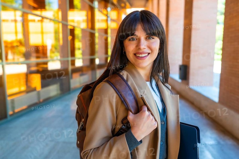Female student standing outdoors on a modern university campus, carrying a backpack and a folder, looking at the camera and smiling, symbolizing education, learning, and academic future