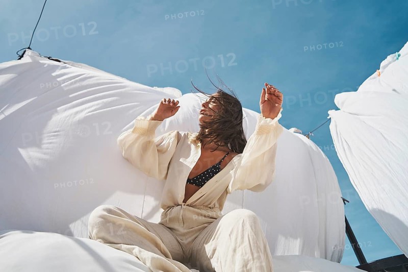A woman is sitting on a rooftop, basking in the sun, surrounded by billowing white fabric, wearing light summer attire The clear blue sky adds a refreshing backdrop