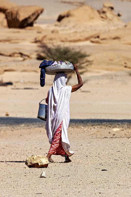 A woman carries loads on her head and a bucket in her hand in a remote desert landscape, woman washing clothes in the Sahara in Algeria