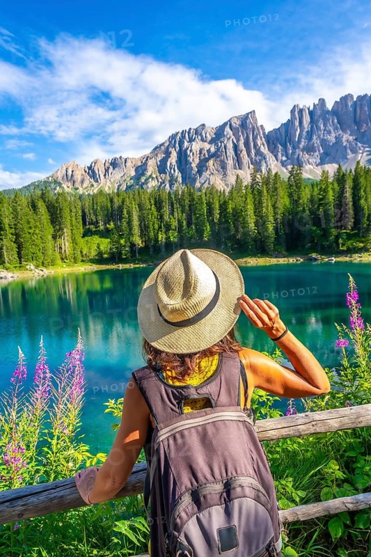 Female tourist with a backpack and hat admiring the turquoise waters of lake carezza, framed by the latemar mountains on a sunny day