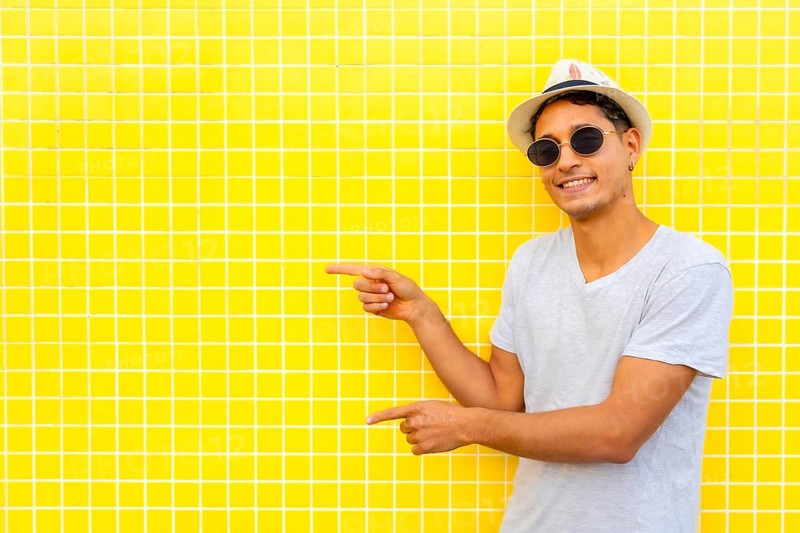 Stylish tourist pointing both index fingers at yellow tiled wall, wearing a hat and sunglasses
