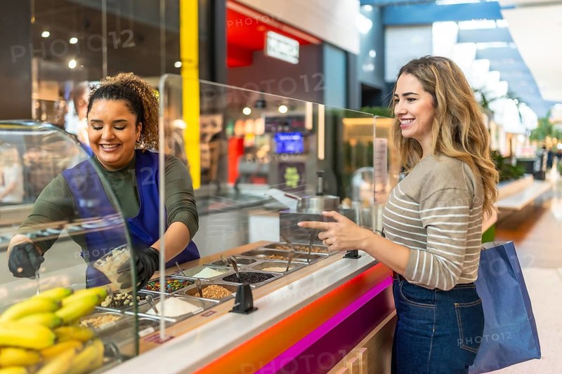 Friendly server preparing a custom dessert for a smiling customer selecting various toppings from a display at a vibrant food kiosk within a shopping mall