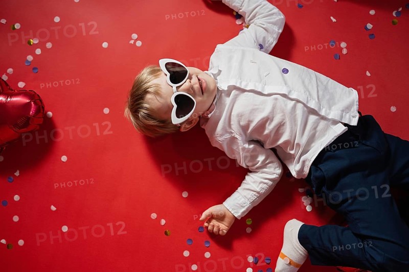 A young boy in heart-shaped sunglasses lies on a red background scattered with colorful confetti and a red heart balloon, capturing the fun and innocence of Valentine's Day