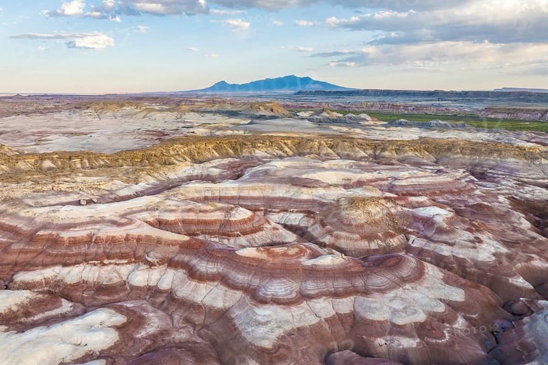 Captivating aerial shot of layered bentonite hills with unique erosion patterns and distant mountains under a wide, cloud-streaked sky, showcasing the geological beauty in Utah, USA