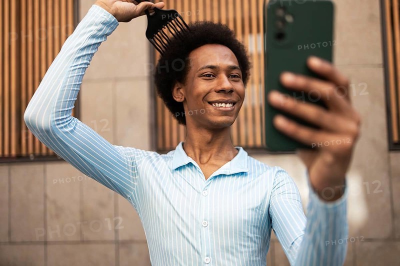 A non-binary individual with an afro hairstyle confidently takes a selfie outdoors. They are smiling, embracing self-expression and authenticity in an urban setting