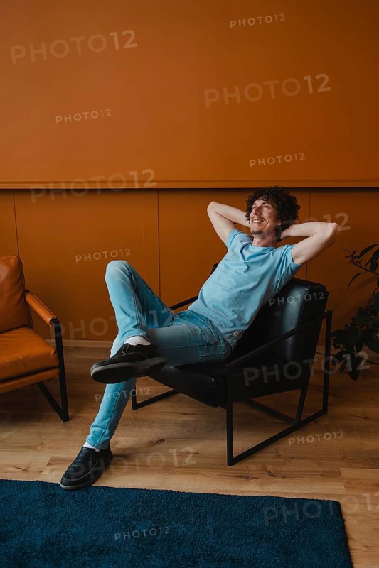 A young man with curly hair relaxes comfortably in a modern chair. His casual attire complements the stylish interior setting, creating a laid-back atmosphere