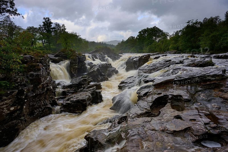 A river with several waterfalls flows through a rocky landscape surrounded by forest under cloudy sky, River Orchy, Eas Urchaidh, Bridge of Orchy, Scotland, United Kingdom, Great Britain
