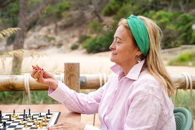 A woman in a pink shirt and green headband holds a chess piece thoughtfully, sitting outdoors at a table with a chessboard A serene background enhances the tranquil moment