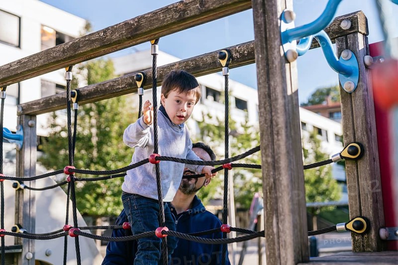 A child with Down syndrome explores a playground climber, supported by his father. The scene is filled with sunlight, showcasing outdoor fun and inclusion