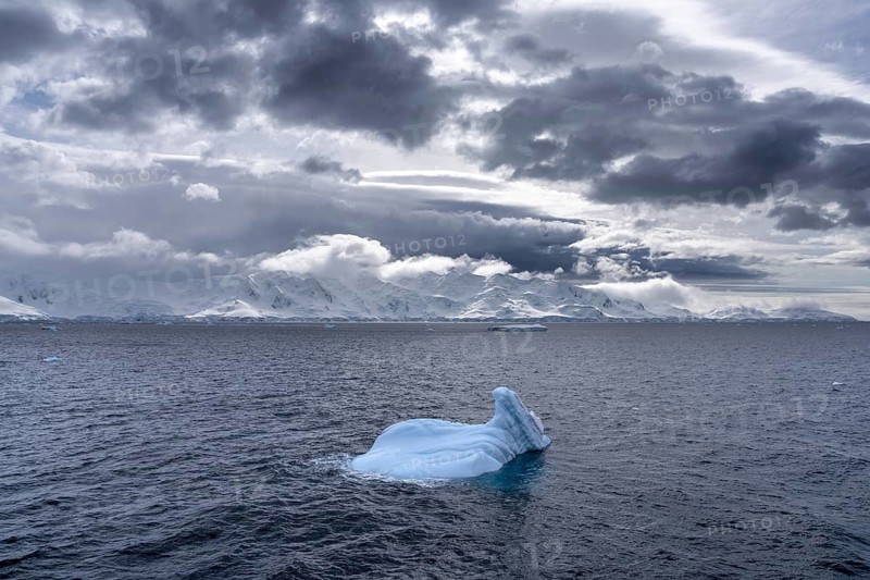 Small iceberg floating in the open water of the Southern Ocean with snow-capped mountains of the Antarctic Peninsula in the background under cloudy skies, Antarctica